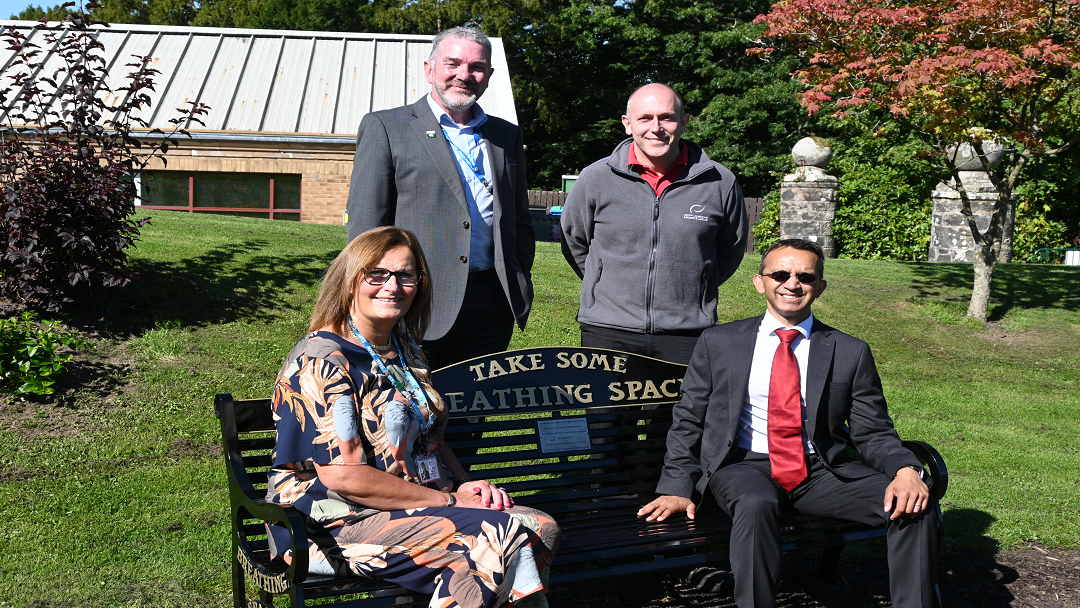 A photo of 4 people, a woman and 3 men, at a bench in a park. The bench says Take Some Breathing Space